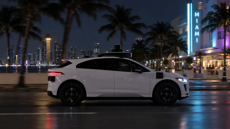 A white robotaxi with roof sensors driving past palm trees and the Miami skyline at night.